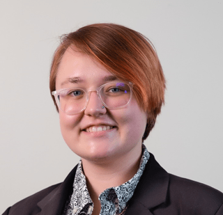 Nina Erbes, a white person with glasses and short reddish-brown hair, smiles at the camera. She wears a black blazer and a black and white button-up shirt.