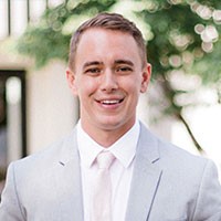 A 30 year old white male with short brown hair is wearing a gray suit with pink tie in front of a soft backdrop featuring the branches of a tree and a building
