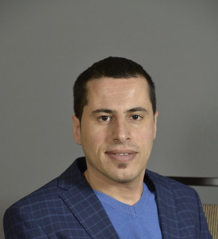 Headshot of Mustafa Rfat, male with dark hair smiling in front of a grey background