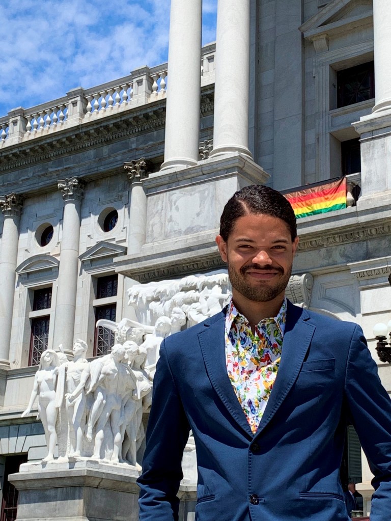 Picture of Parris, standing in front of the Pennsylvania State Building. He is looking at the camera, smiling, and wearing a floral dress shirt with a blue sport coat.