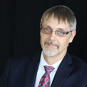 Image of John Tschida a while man with glasses greying hair and mustache wearing a black suit and red tie with white shirt smiling in front of black background.