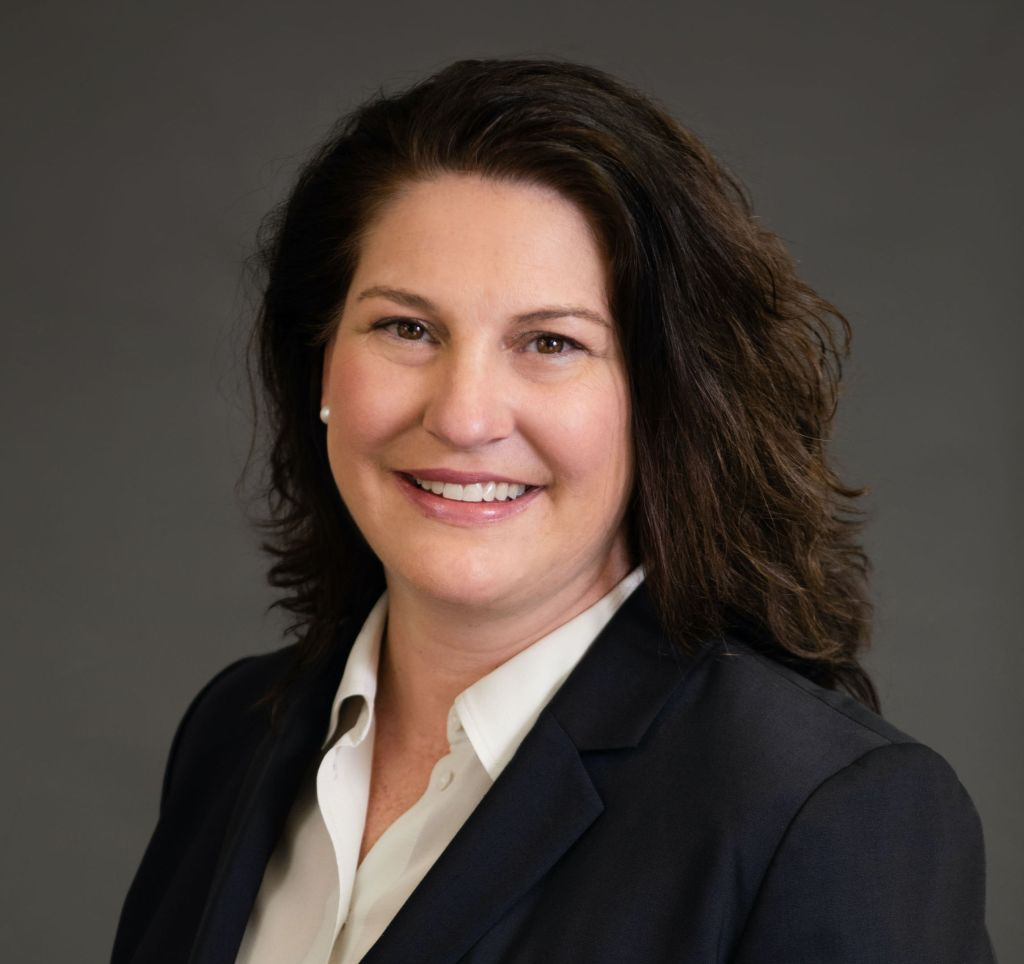 Image of Sara Bitter a white woman with dark brown hair in a white collared blouse and black blazer smiling in front of a grey background