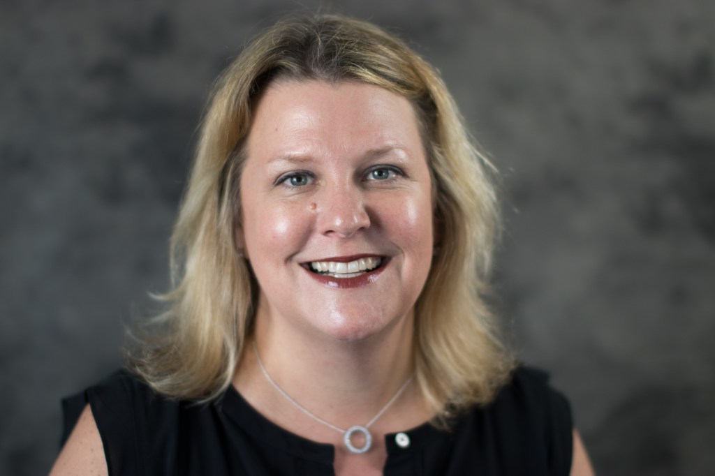 Maureen a white woman with blond hair wearing a black top smiling with red lipstick in front of a grey backdrop