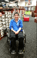 Image of Agatha Gietzen. White woman in wheel chair blue shit and black pants smiling in front of shoe aisle. 