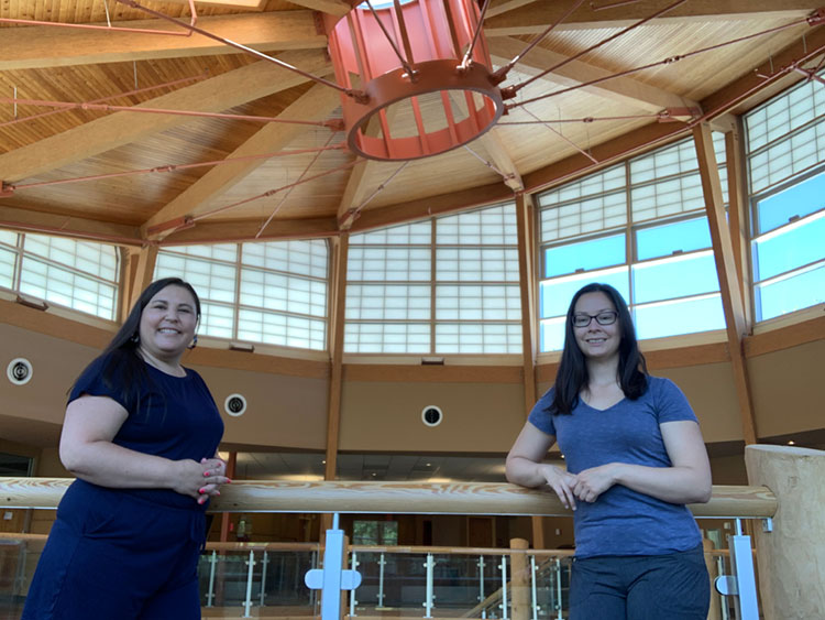 Salena standing in a blue dress and Helen standing in a blue t-shirt and jeans. The background is a large room with a wooden ceiling. 