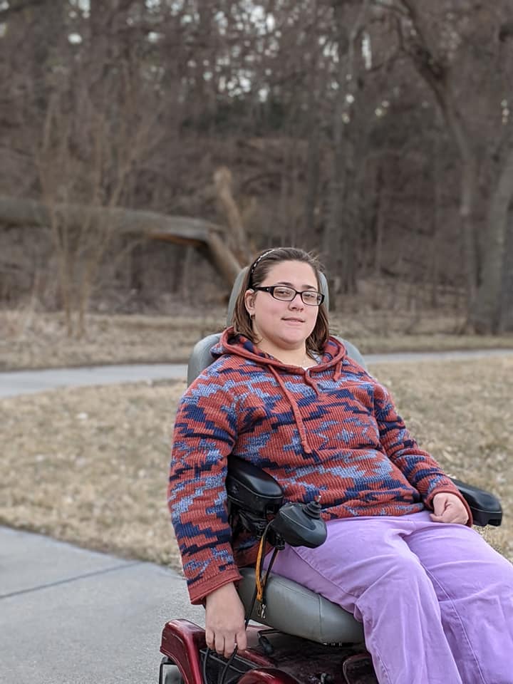 the author, Melanie Davis, wearing a multicolored sweatshirt and purple pants, sitting in a motorized red wheelchair outside. 