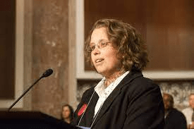 White woman with light curly hair and glasses, speaking at podium