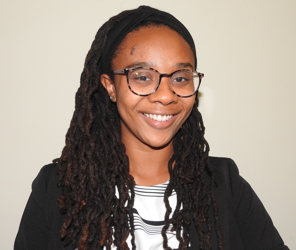 Young woman with glasses and long hair, wearing a black jacket and striped shirt underneath.