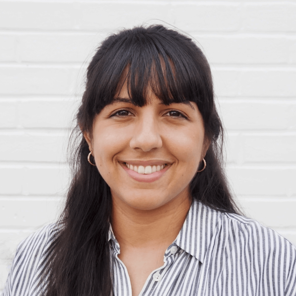 Lilian Aluri smiles at the camera in front of a white brick wall. She has medium-brown skin and long dark brown hair with banks. She wears a white and blue striped button down shirt with gold hoop earrings.
