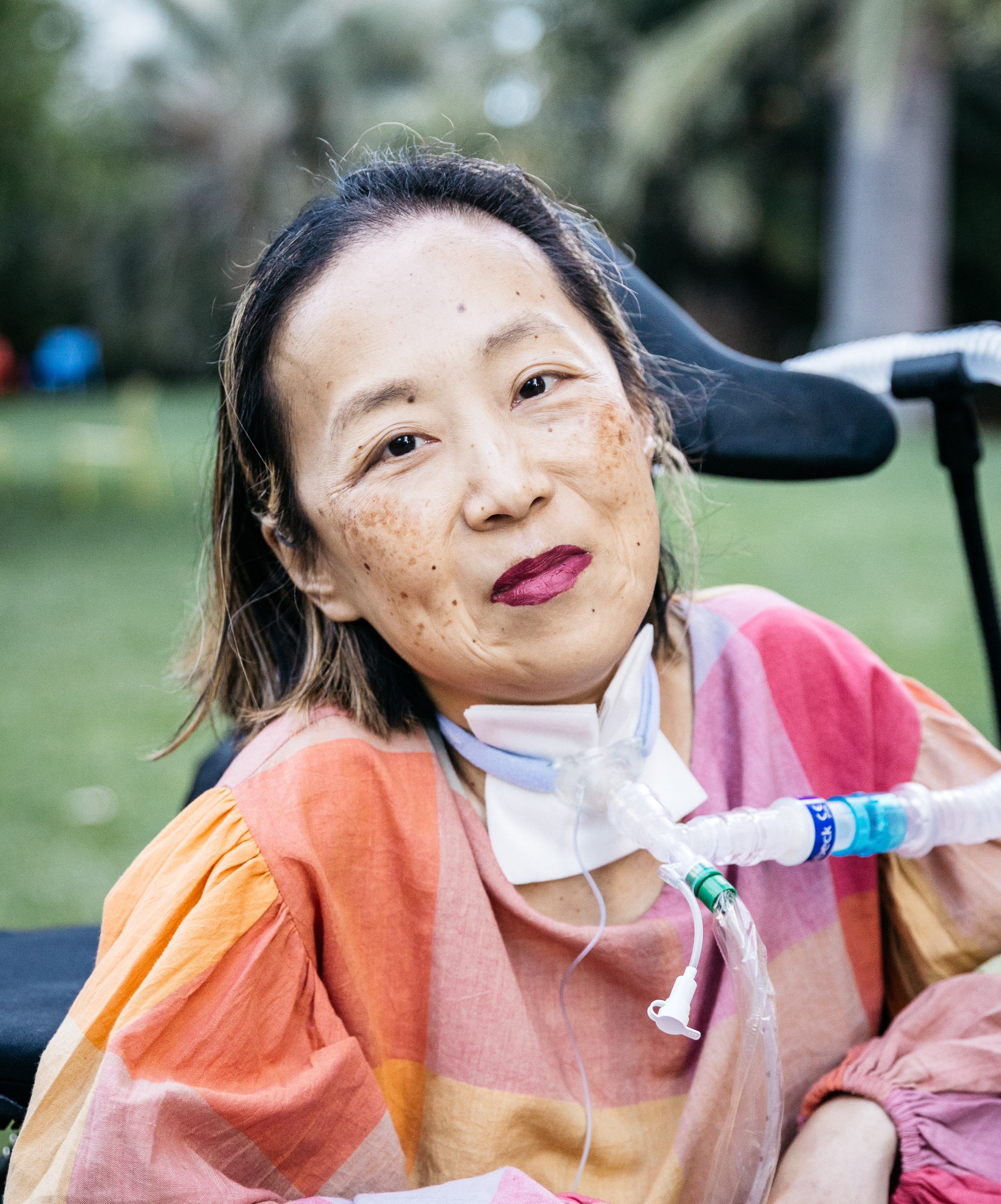 The photo shows an adult Asian woman sitting outdoors, possibly in a park. She has shoulder-length hair and is wearing a colorful, loose-fitting top with warm tones like orange, pink, and yellow. She appears to be using a wheelchair and has a tracheostomy tube connected to respiratory equipment. Her expression is calm and composed, and she is wearing dark lipstick. The background is filled with greenery and blurred trees, giving the scene a peaceful, natural atmosphere.